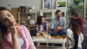Young woman sitting apart from a group gathering, looking thoughtful and disconnected while others socialise in the background