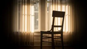 Empty wooden chair by a sunlit window with soft curtains, symbolising stillness, reflection, rest, and emotional space